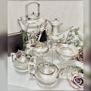May include: A complete silver tea set, including a teapot, coffee pot, creamer, sugar bowl, and serving tray. The set has a reflective surface and is arranged on a white surface with floral accents. The tea set is a classic design.