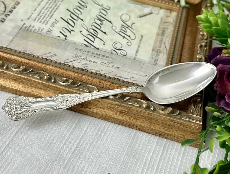 May include: A polished silver-toned spoon featuring an intricate embossed floral and shell pattern along the handle. The spoon has a smooth, oval-shaped bowl and a decorative, ornate design extending from the base of the handle to the neck.