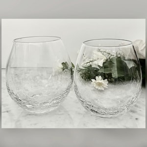 May include: Two clear, stemless wine glasses with a crackled texture. One glass is empty, and the other contains white flowers and greenery. The glasses are on a white marble surface.