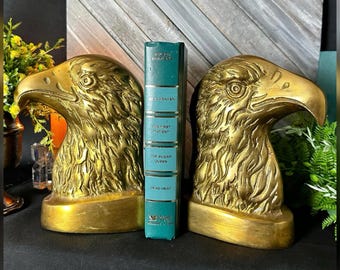 May include: A pair of gold-toned metal eagle head bookends with detailed feather textures, positioned on either side of a single teal-colored book with gold lettering on the spine that reads "Blue Heaven," "The First Patient," "The Sugar Queen," and "Dead Heat."