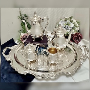 May include: A silver tea set with a decorative tray. The set includes a teapot, a creamer, a sugar bowl, and a serving tray. The tray has ornate edges and a floral design. The teapot is on a small stand. The set is displayed with books and flowers.