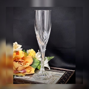 May include: A clear glass champagne flute with a cut crystal design. The flute is sitting on a decorative surface with a floral arrangement.
