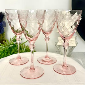 May include: A set of four pink glass goblets featuring a textured diamond pattern on the bowl and a delicate etched lace-like band near the rim. Each glass has a slender stem with a tiered base and a wide, circular foot for stability.