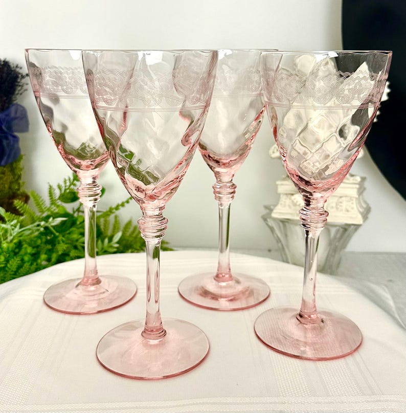 May include: A set of four pink glass goblets featuring a textured diamond pattern on the bowl and a delicate etched lace-like band near the rim. Each glass has a slender stem with a tiered base and a wide, circular foot for stability.