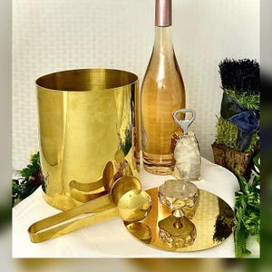 May include: A collection of gold-toned barware and serving accessories. Includes a cylindrical ice bucket, a wine bottle, a bottle opener, serving tongs, and a decorative cake stand. The items are arranged on a white surface with greenery accents.