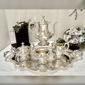 May include: A silver tea set with ornate floral detailing, including a teapot, sugar bowl, creamer, and a small bowl, arranged on a matching oval tray. The set is displayed on a white surface, with floral arrangements in the background.