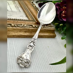 May include: A silver spoon with an ornate floral design on the handle. The spoon has a large, oval bowl and a detailed pattern. The handle is engraved with initials. The spoon is likely vintage or antique.