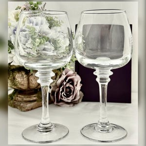 May include: Two clear wine glasses with a decorative stem and a round base. The glasses are empty, and the background features a floral arrangement with white and purple flowers and a large rose.