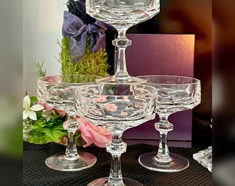 May include: A set of four clear crystal coupe glasses featuring a wide, shallow bowl with a geometric faceted pattern. Each glass has a short, multi-faceted stem and a circular base, arranged in a tiered formation to showcase the intricate glasswork.