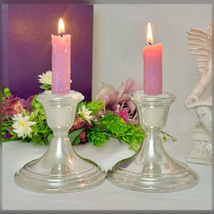 May include: Two silver candlesticks each hold a lit pink candle. The candles are dripping wax. The candlesticks are on a white surface, with flowers and a purple book in the background.