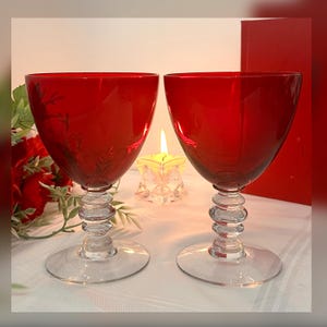 May include: Two ruby red wine glasses with clear stems and bases. The bowls are a deep red, and the stems have a stacked, rounded design. A lit candle and red roses are in the background.