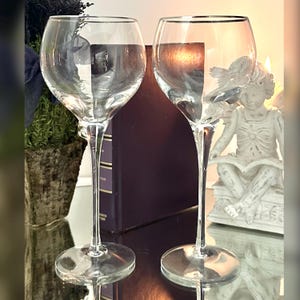 May include: Two clear wine glasses with long stems and rounded bowls. The glasses have a silver rim and are filled with a clear liquid. The glasses are on a reflective surface, with a small statue and a book in the background.