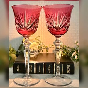 May include: Two matching wine glasses with deep red bowls featuring a cut glass starburst pattern. The stems are clear, faceted glass with a multi-sided design leading to a circular base. The glasses are positioned in front of a black book.