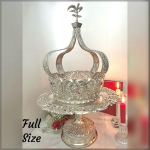 May include: Ornate silver-colored crown-shaped centerpiece with intricate detailing, set on a decorative pedestal. The crown features a fleur-de-lis finial and is adorned with red and blue accents. The text "Full Size" is visible on the left side.