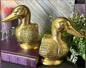 May include: A pair of polished brass duck bookends, each featuring detailed feather engravings and a smooth, reflective finish. The ducks are depicted in a profile view with heads turned, showcasing a classic, solid metal construction.
