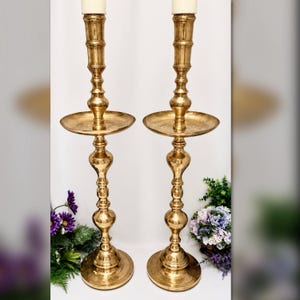 May include: A pair of tall, polished brass candlesticks featuring a tiered, turned design with a wide circular drip tray and a stable, round base. Each candlestick holds a single cream-colored taper candle, showcasing a metallic finish.