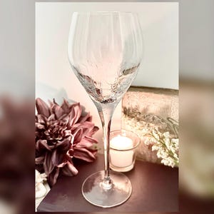 May include: A tall, clear wine glass with a textured design on the upper portion. The glass has a long stem and a round base. The image also includes a dark pink flower, a candle, and a decorative box.