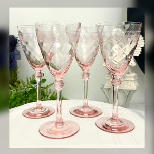 May include: Four vintage pink-tinted wine glasses. The glasses have a delicate, etched design around the rim and a textured pattern on the bowl. The stems and bases are also pink. The glasses are arranged on a white surface.