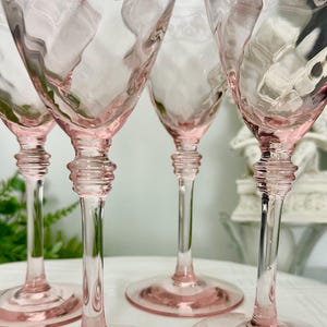 May include: A set of four light pink glass goblets featuring a textured diamond pattern on the bowl, a clear stem with a multi-ringed decorative detail, and a wide circular base. The upper rim of each glass displays a delicate etched floral design.