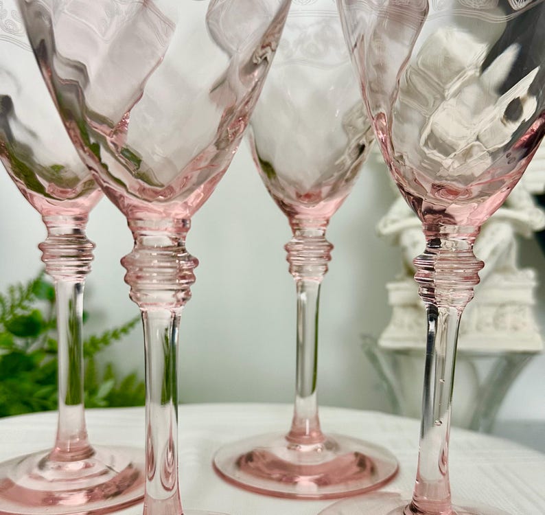 May include: A set of four light pink glass goblets featuring a textured diamond pattern on the bowl, a clear stem with a multi-ringed decorative detail, and a wide circular base. The upper rim of each glass displays a delicate etched floral design.
