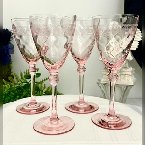 May include: A set of four light pink glass wine glasses featuring a wavy rim, a textured swirl pattern on the bowl, and an etched lace-like band near the top. Each glass has a slender stem with a decorative ring detail and a circular base.