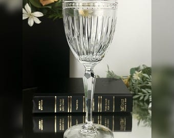 Könnte beinhalten: Ein klares Kristall-Weinglas mit vertikalen geätzten Linien. Das Glas hat einen langen Stiel mit Facettendesign und einen runden Fuß. Der Hintergrund umfasst Bücher und Blumengestecke.
