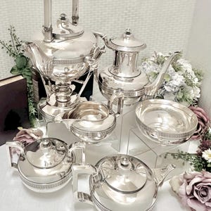 May include: A complete silver tea set, including a teapot with a warming stand, a coffee pot, two teapots, a creamer, and a sugar bowl. The set features ornate detailing and a polished finish, perfect for elegant tea parties.