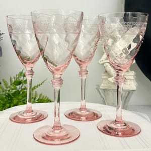 May include: A set of four light pink glass wine glasses featuring a textured, wavy bowl surface and a delicate etched lace-like pattern around the upper rim. Each glass has a slender stem with a tiered base and a wide, circular foot.