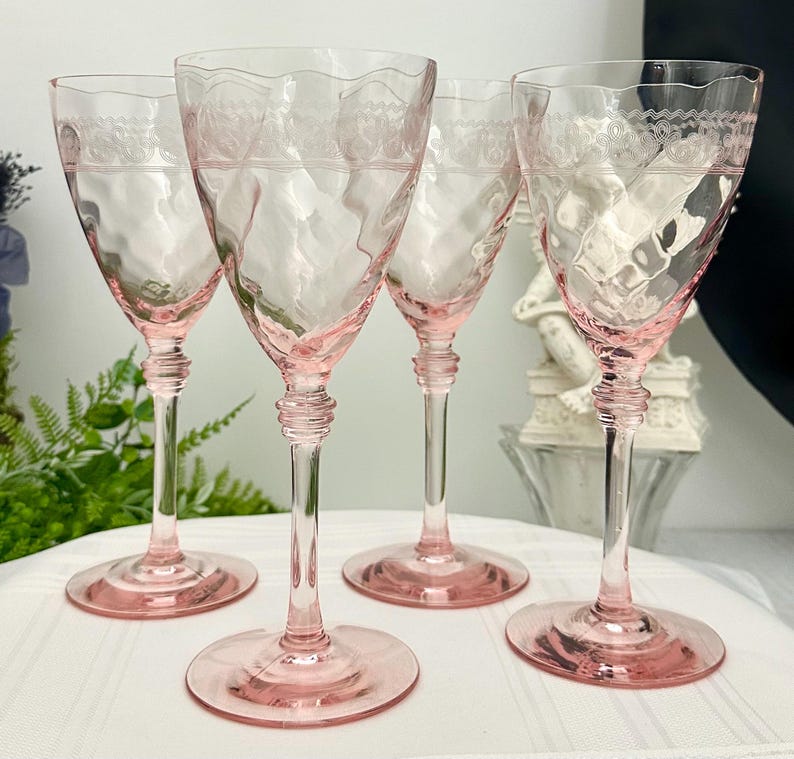 May include: A set of four light pink glass wine glasses featuring a textured, wavy bowl surface and a delicate etched lace-like pattern around the upper rim. Each glass has a slender stem with a tiered base and a wide, circular foot.