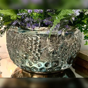 May include: A round, metallic silver-toned bowl featuring a raised, embossed relief pattern of grape clusters and vine leaves. The bowl is filled with an arrangement of green foliage and small, light purple flowers.