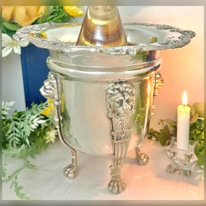 May include: A silver-colored metal ice bucket with lion head details and claw feet. The bucket has a decorative rim and holds a clear glass bottle. A lit white candle sits nearby in a decorative holder.