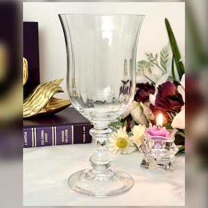 May include: A clear, stemmed glass with a wide, flared bowl. The glass sits on a round base and has a decorative stem. A lit candle in a star-shaped holder and a book are in the background. The glass is likely for serving beverages.