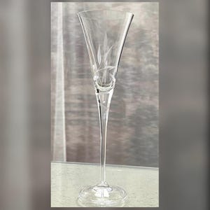 May include: A tall, clear glass champagne flute featuring a slender stem and a flared bowl. The glass is decorated with a delicate, etched leaf pattern that wraps around the upper portion of the bowl, providing a subtle, textured visual detail.