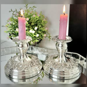 May include: Two silver-toned metal candle holders with a wide, circular base featuring an embossed floral pattern. Each holder supports a single pink taper candle with a lit flame, showing visible wax drips along the sides of the candles.