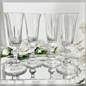 May include: Four clear, stemmed champagne flutes with vertical etched lines on the bowl. The flutes sit on a mirrored tray with a decorative edge. The stems have a detailed, bulbous design. White flowers are visible in the background.