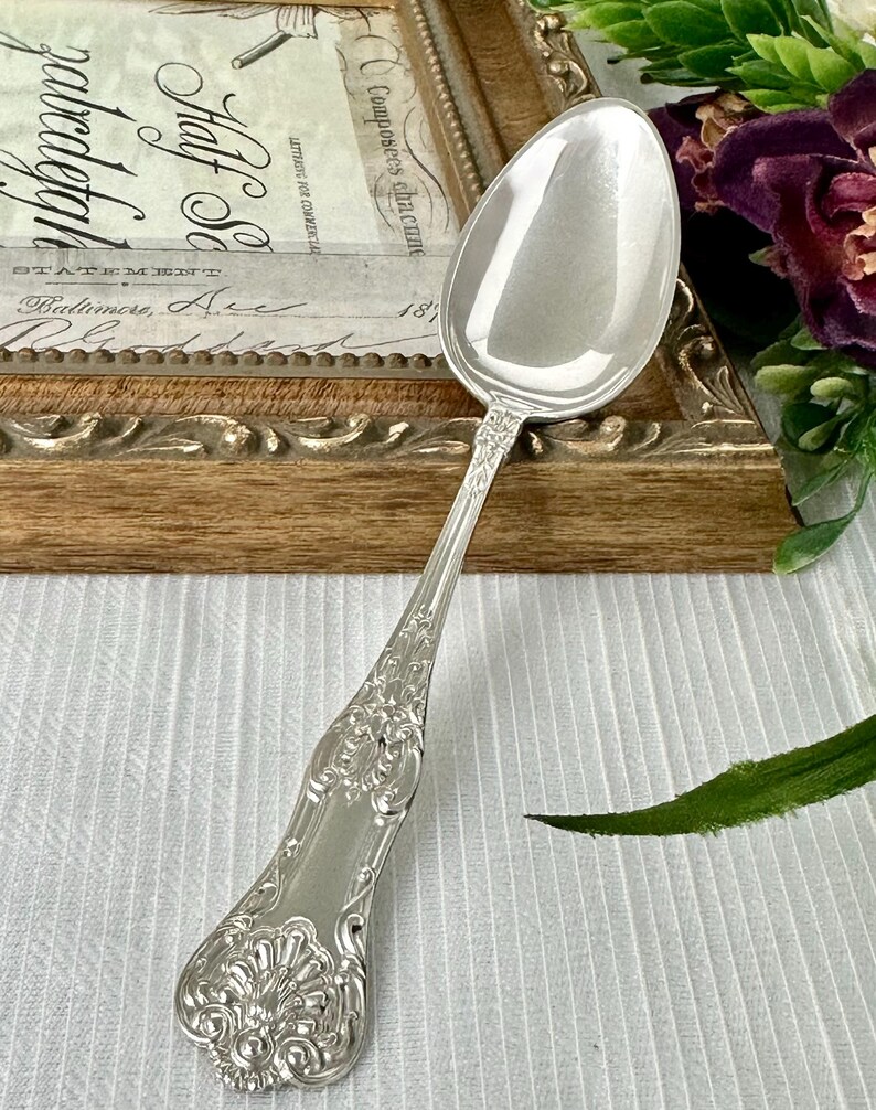 May include: A polished silver-tone metal spoon featuring an ornate, embossed floral and scrollwork pattern along the handle. The spoon has a smooth, reflective oval bowl and a decorative, flared end with intricate detailing on the handle surface.