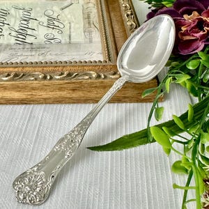 May include: A polished silver-toned spoon featuring an ornate, embossed floral and shell pattern along the handle. The spoon has a smooth, reflective oval bowl and a decorative, flared end, showcasing intricate metalwork details throughout the handle.