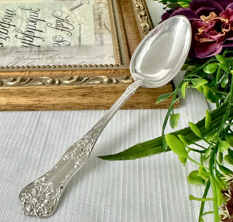 May include: A polished silver-toned spoon featuring an ornate, embossed floral and shell pattern along the handle. The spoon has a smooth, reflective oval bowl and a decorative, flared end, showcasing intricate metalwork details throughout the handle.