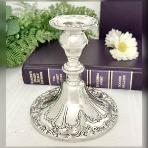 May include: A silver candlestick with an ornate, detailed design. The base is wide and flared, with a decorative edge. The stem is a classic shape, and the top has a small, circular holder for a candle. A classic home decor item.
