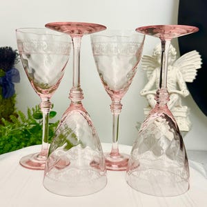 May include: Four pink glass goblets with tall stems and etched lace-like patterns around the upper bowls. Two glasses stand upright while two are inverted, showcasing the delicate glasswork and the translucent pink hue of the stemware set.