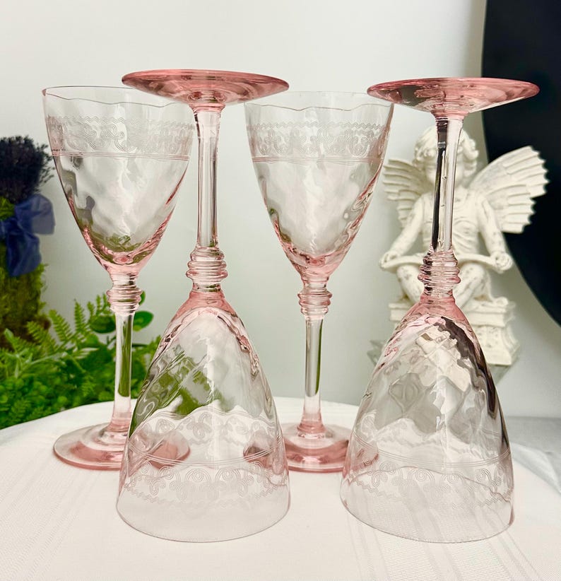 May include: Four pink glass goblets with tall stems and etched lace-like patterns around the upper bowls. Two glasses stand upright while two are inverted, showcasing the delicate glasswork and the translucent pink hue of the stemware set.