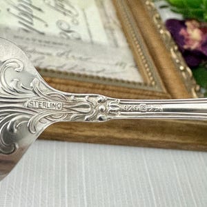 May include: A silver spoon featuring an ornate, embossed scrollwork pattern on the handle. The metal surface is stamped with the word STERLING and the number 925, indicating the composition of the silver material used in this flatware piece.