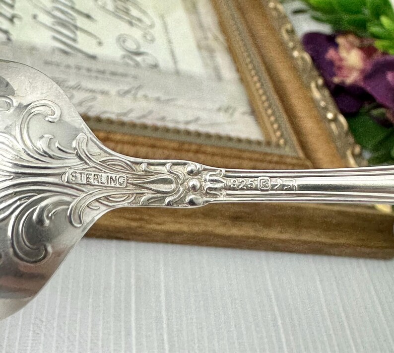 May include: A silver spoon featuring an ornate, embossed scrollwork pattern on the handle. The metal surface is stamped with the word STERLING and the number 925, indicating the composition of the silver material used in this flatware piece.