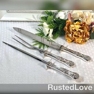 May include: A set of three silver-plated serving utensils, including a carving knife, a fork, and a serving knife. The utensils have ornate handles with a floral design.