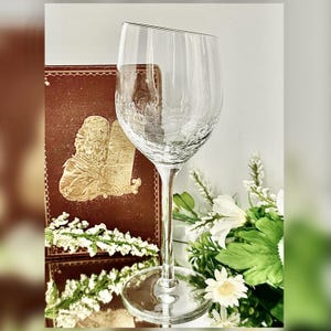 May include: A clear wine glass with a textured bowl and a long stem. The glass is set against a backdrop of a decorative book and white flowers. The glass has a unique, angled rim.