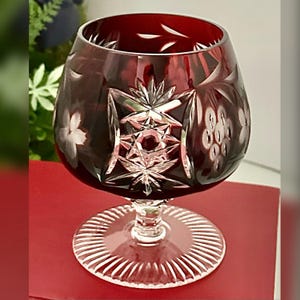 May include: A ruby red crystal brandy snifter with intricate cut glass designs. The bowl features starburst and floral patterns, with a clear, ribbed base. The glass is set against a red background, highlighting its rich color and craftsmanship.