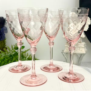 May include: A set of four light pink glass wine glasses featuring a wavy rim, a textured hammered bowl, and an etched decorative band near the top. Each glass has a slender stem with a ringed detail above a circular base, standing upright.