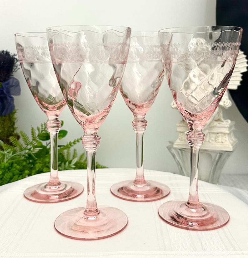 May include: A set of four light pink glass wine glasses featuring a wavy rim, a textured hammered bowl, and an etched decorative band near the top. Each glass has a slender stem with a ringed detail above a circular base, standing upright.