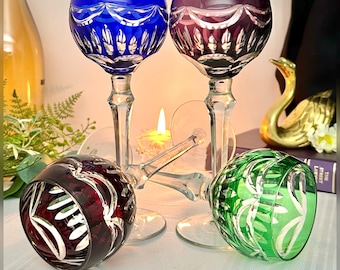 May include: Four cut-to-clear crystal wine glasses featuring intricate etched patterns. Two glasses stand upright in blue and purple, while two glasses lie horizontally in red and green. Each glass has a clear faceted stem and base.