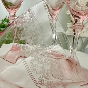 May include: A set of light pink glass goblets featuring a delicate etched pattern of swirling lines and geometric zigzags. The glassware has a clear stem and a wide, flared bowl, with the etched design prominently displayed on the surface.